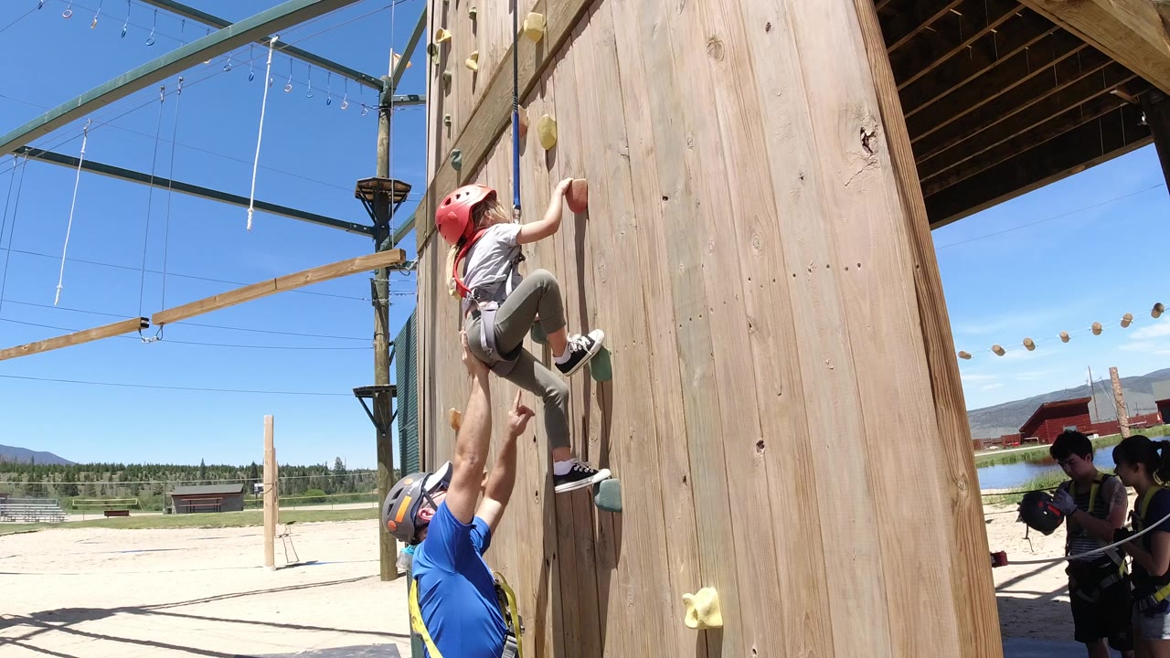 In Winter Park, Colorado, an adult in a blue shirt reaches up to support a helmeted child starting up a wooden climbing wall, with ropes-course beams and blue sky behind them.