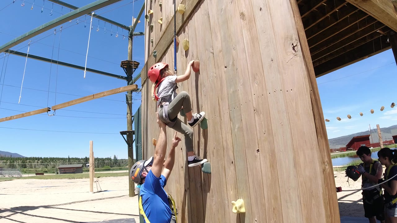 The child is slightly higher on the wooden climbing wall in Winter Park, Colorado, while the adult keeps one arm raised and other harnessed people stand near the rope area in the background.
