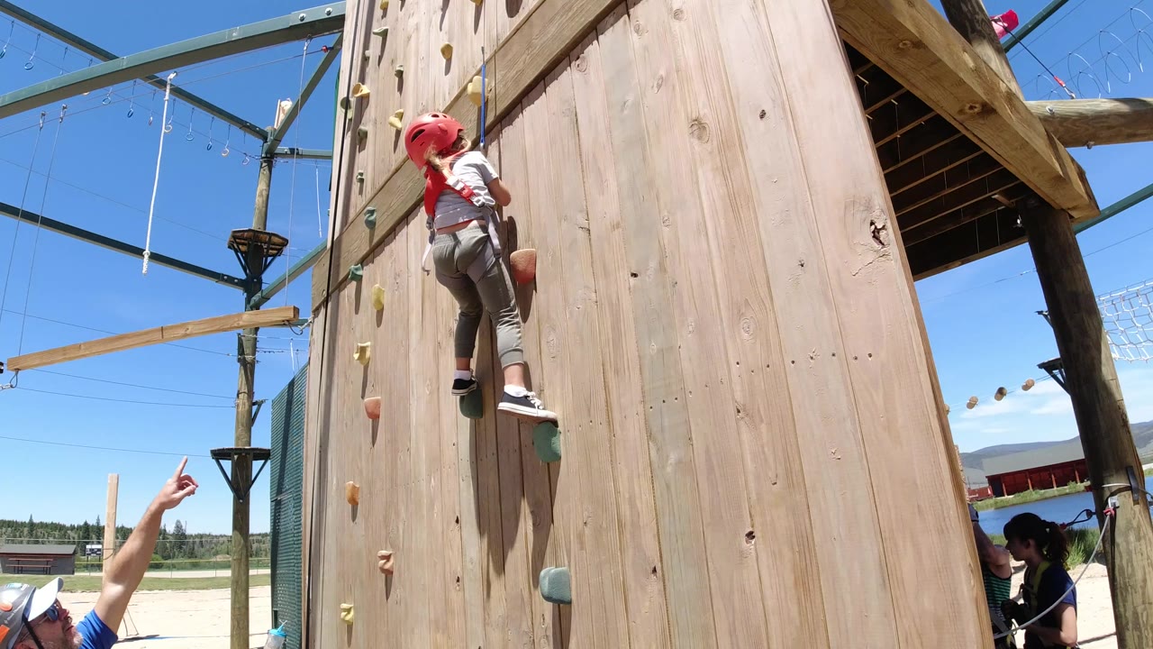 The harnessed child continues upward on the wooden climbing wall in Winter Park, Colorado, with colored holds, safety rope, a partial adult figure, and blue sky around the course framing.