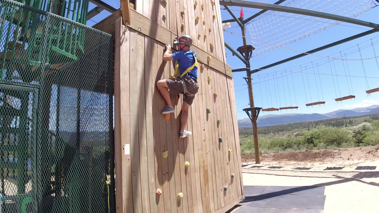 Man in a blue shirt climbs a wooden wall beside green metal stairs.