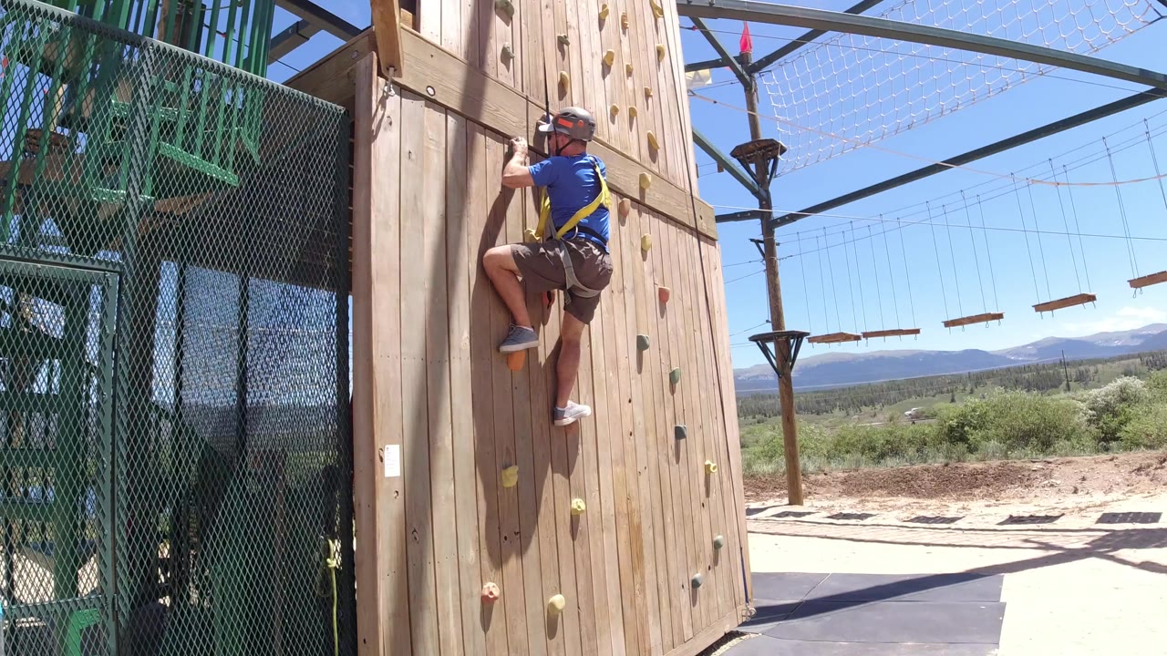 The harnessed climber stays on the wooden wall in Winter Park, Colorado, with colored holds in front of him and suspended ropes course elements visible across the sky.