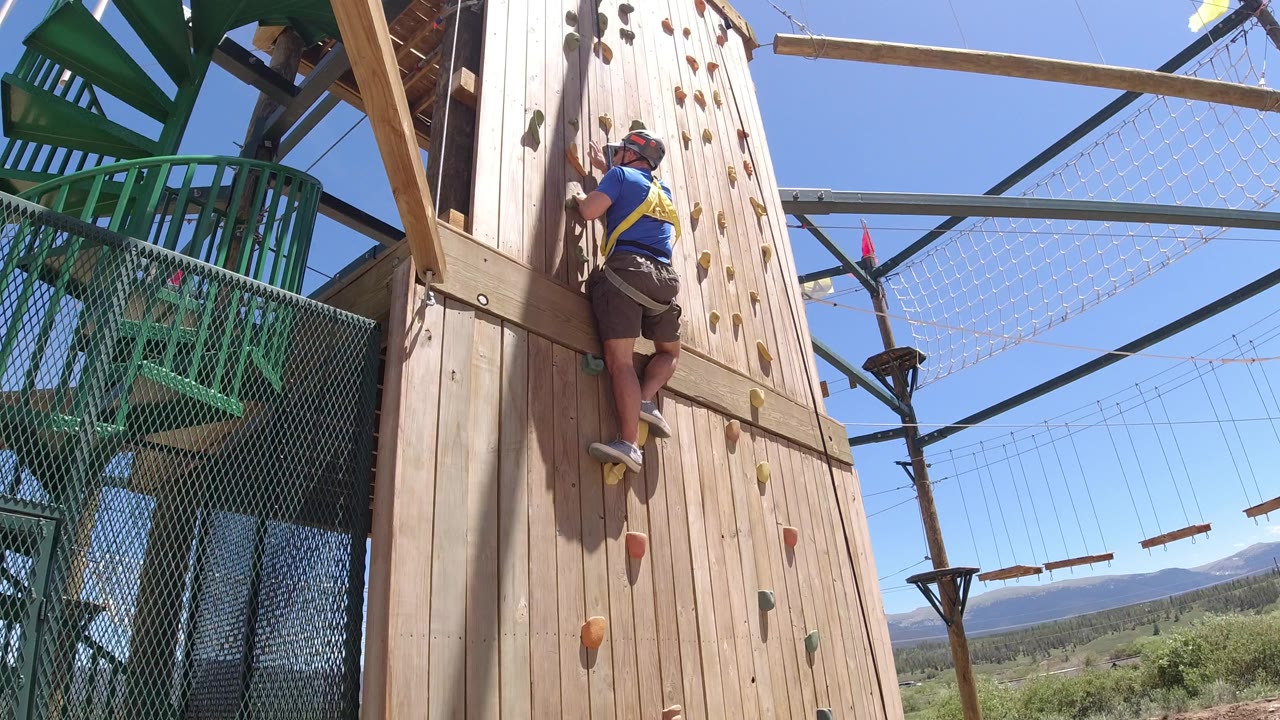 Helmeted man climbing a wooden artificial wall beside green ropes-course platforms.