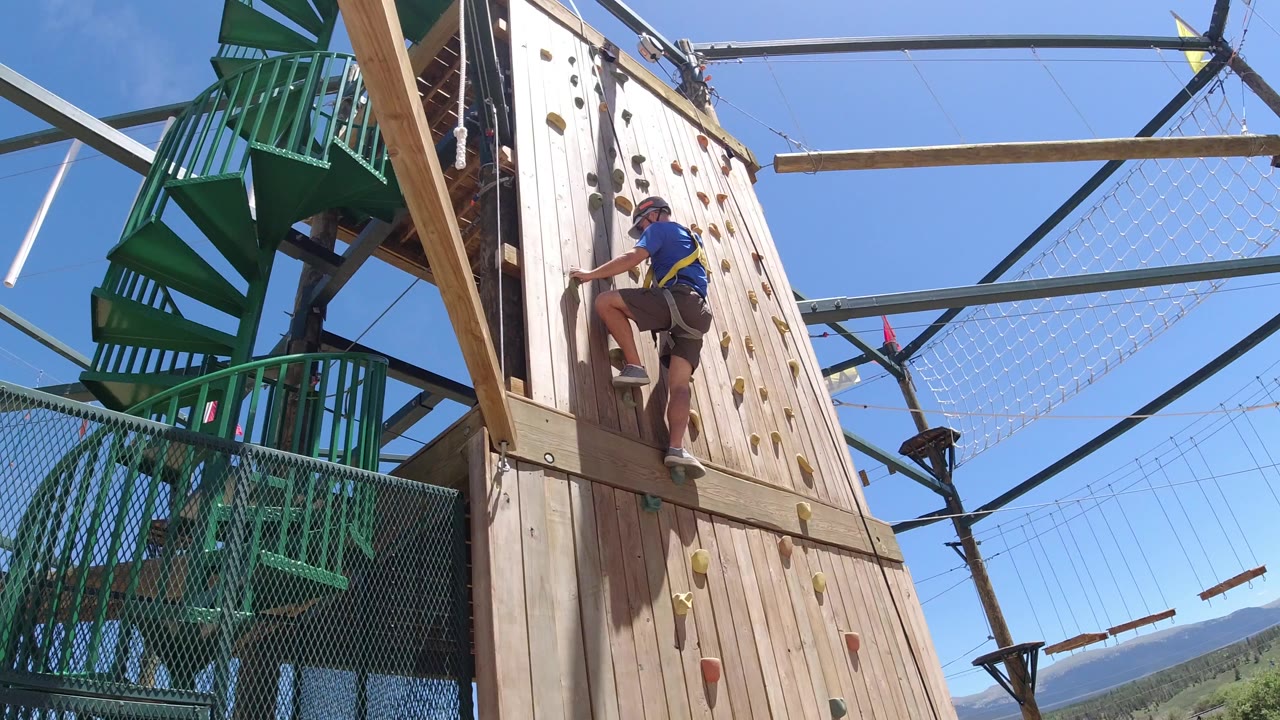 Person climbing a wood rock wall beside green stairs and ropes.