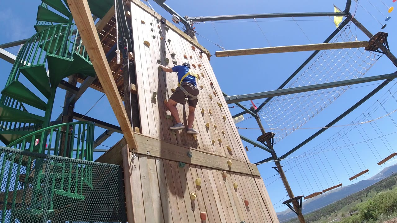 A child in a blue shirt and harness climbs the left side of a tall wooden wall at an outdoor recreation course in Winter Park, Colorado, with green stairs and rope obstacles behind.