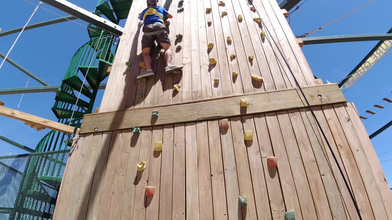 Man climbing a wooden outdoor wall with colored holds.