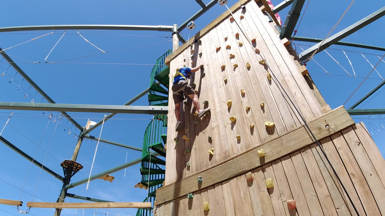 The young climber stays near the upper left of the Winter Park climbing wall, with yellow and orange holds running up the sunlit boards.