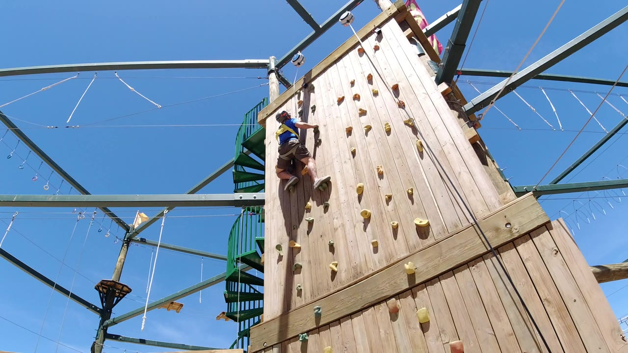 At 0:00, a child climbs the left side of a wooden rock wall in Winter Park, Colorado, with colored holds, green metal beams, cables, and blue sky visible.