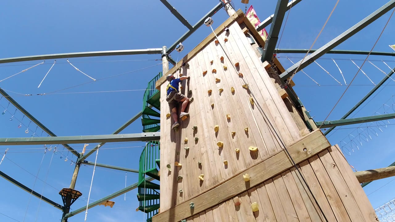 Helmeted man climbing a tall wooden wall under a blue sky.