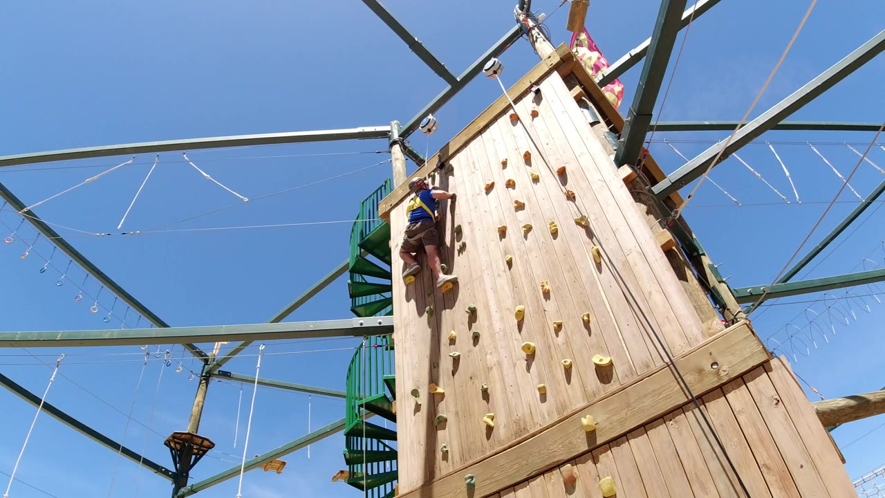 In Winter Park, Colorado, a helmeted climber is partway up the left side of a tall wooden climbing wall under green steel beams and clear blue sky.