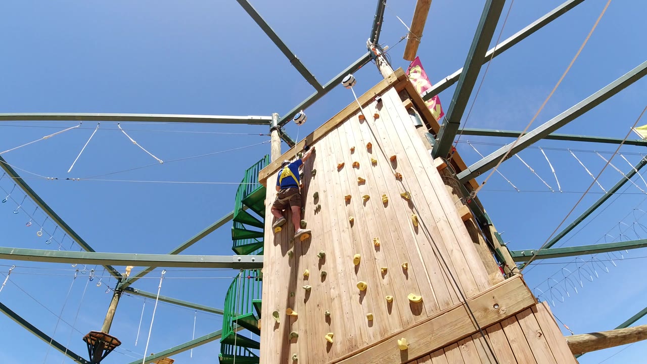 At 0:01 in Winter Park, Colorado, the climber remains on the left side of the wooden wall while overhead beams and ropes stretch across the bright sky.