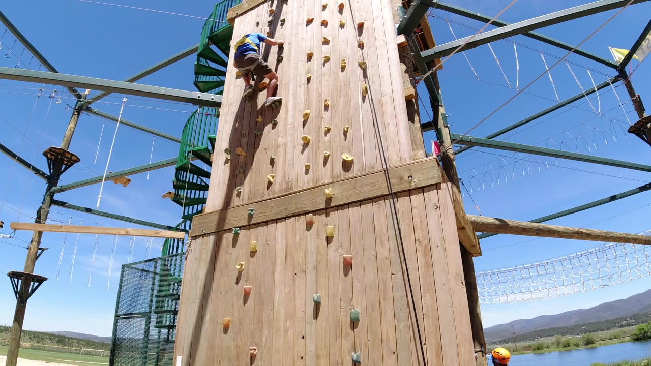 At 0:00 in Winter Park, Colorado, a helmeted climber is near the upper left of a tall wooden climbing wall beneath green metal course framing and a clear blue sky.