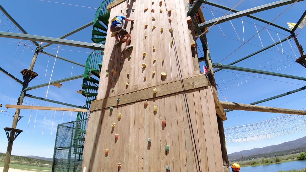 At 0:01 in Winter Park, Colorado, the climber remains on the upper left side of the wooden wall, with colored holds, overhead beams, suspended ropes, and distant hills visible beyond the structure.