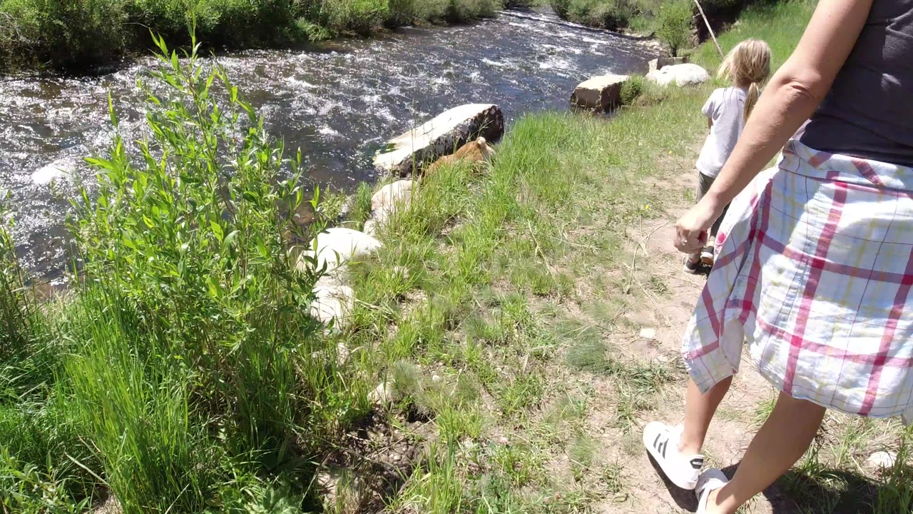 At 0:01, the woman remains cropped close at right while the child moves ahead near the river, with whitewater, green plants, and pale rocks visible in Winter Park, Colorado.