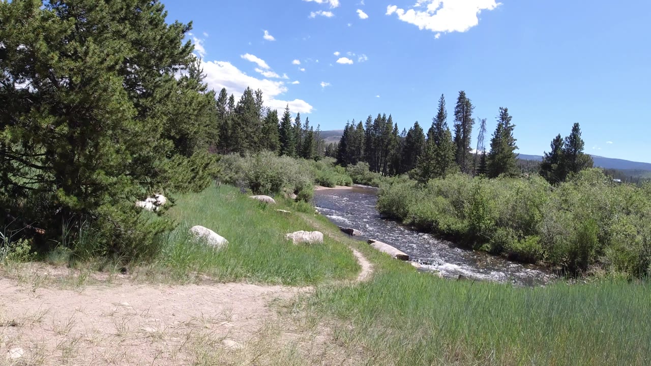 Dirt path curves beside a fast stream and grassy riverbank.