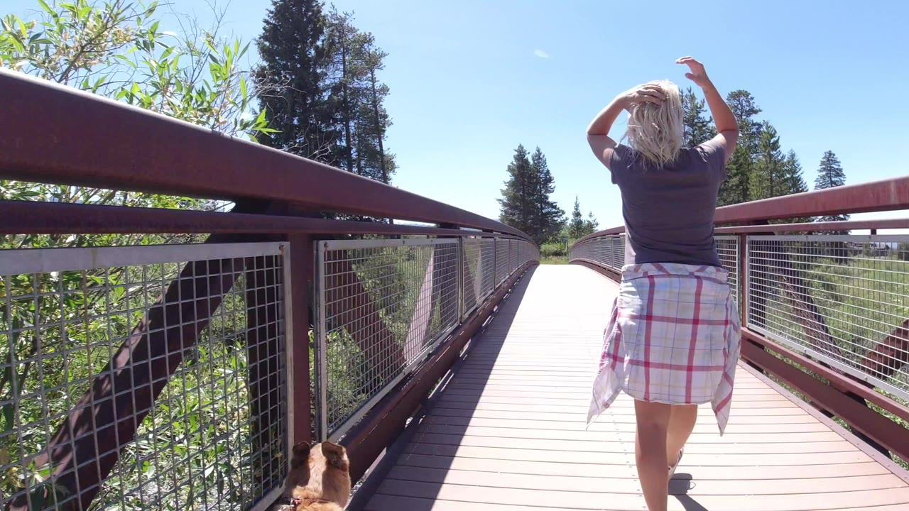 A blonde woman walks away along a red metal pedestrian bridge in Winter Park, Colorado, with a dog partly visible at the lower left edge and trees beyond the railings.