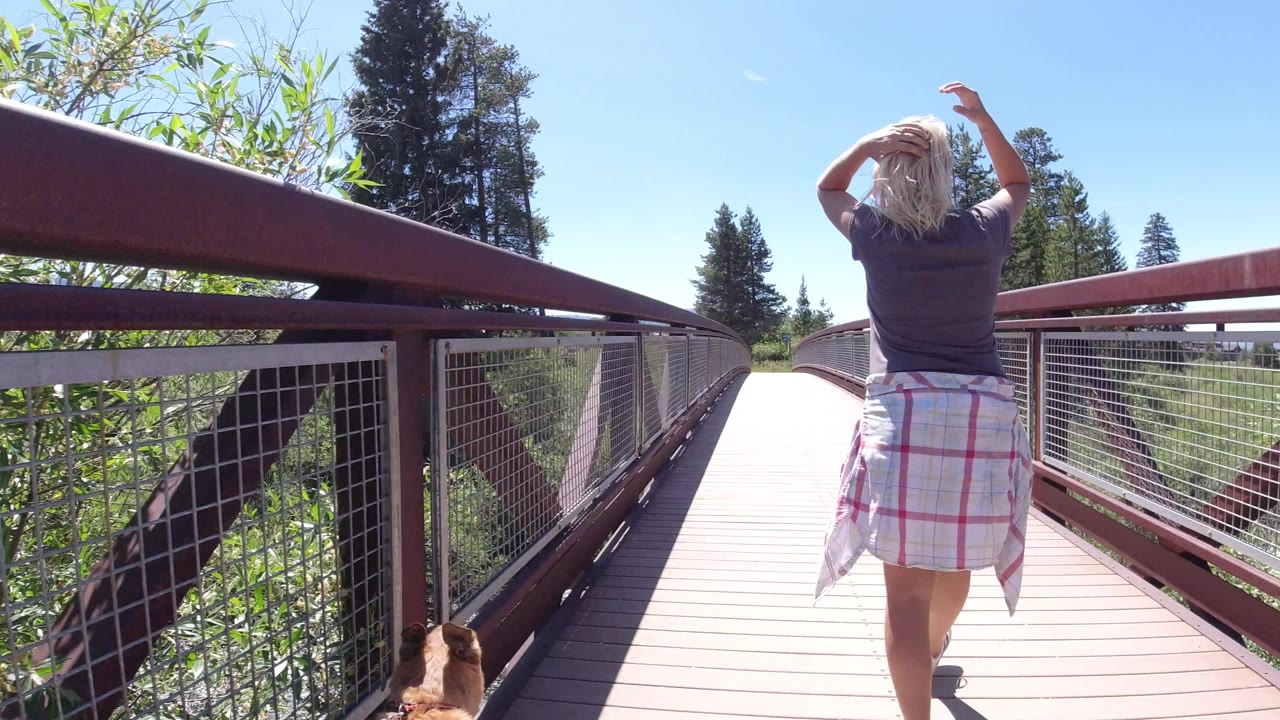 The woman remains centered on the Winter Park pedestrian bridge, raising her hands toward her hair while the dog is partly visible near the lower frame edge.