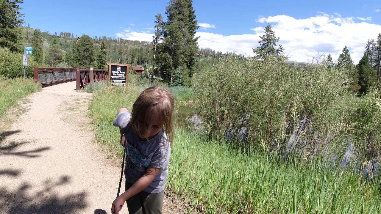 The young girl remains near the path edge in Winter Park, Colorado, bending forward with the stick while the red bridge and trail sign sit farther up the path.