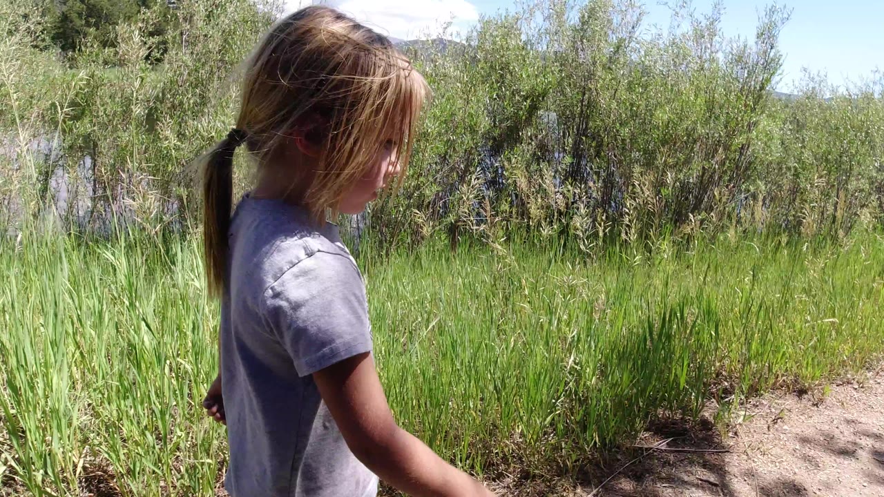 The girl continues along the path in Winter Park, Colorado, seen from the side with windblown hair, green roadside vegetation, and distant snow visible above the shrubs.
