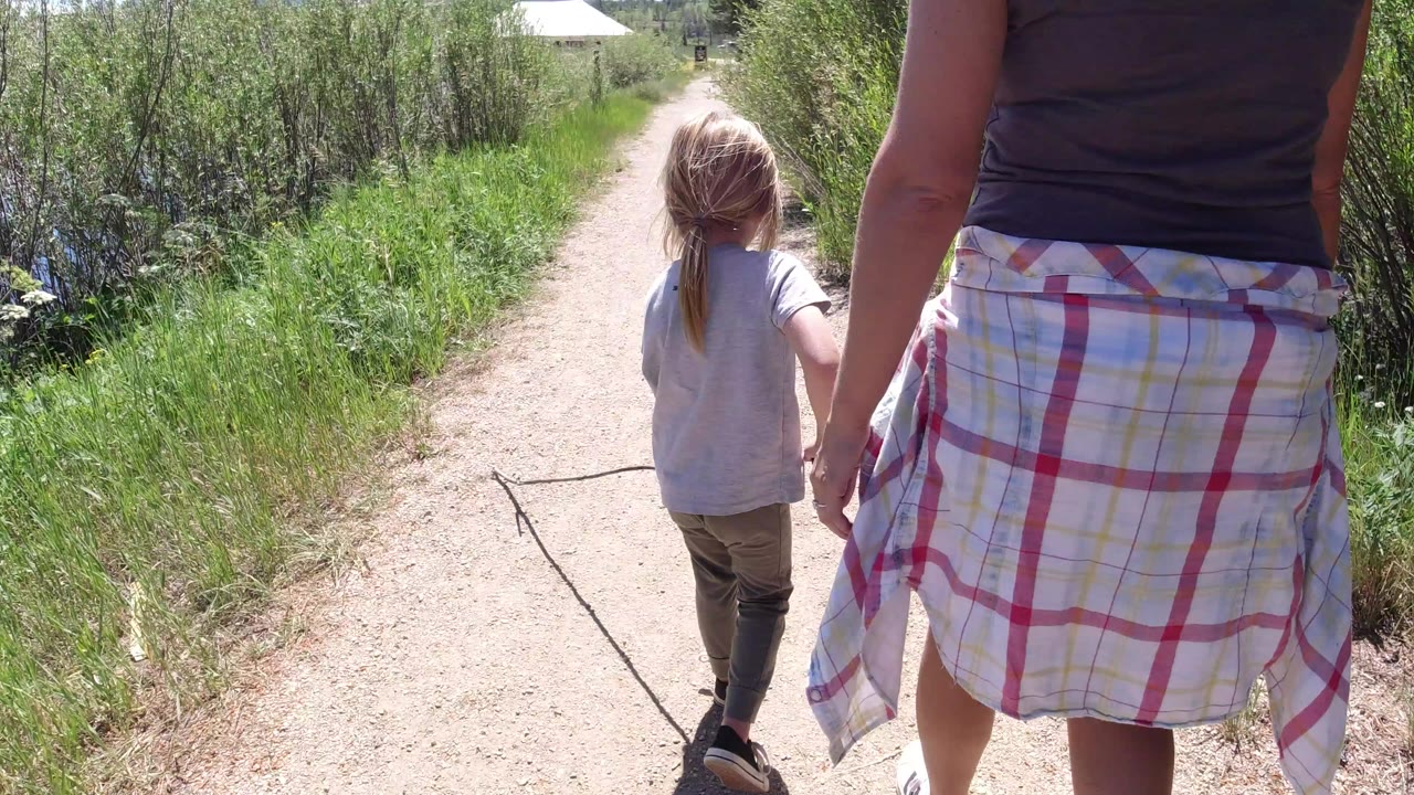 A child and an adult walk away on a dirt path in Winter Park, Colorado, with bright sun on tall grass and brush along both sides.