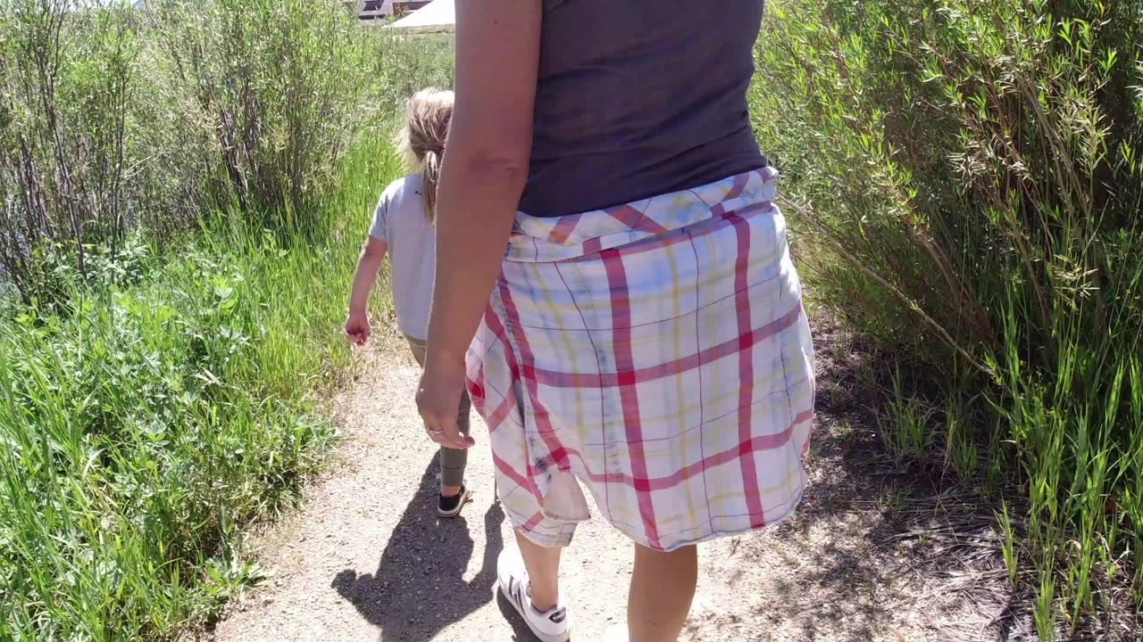 An adult and child walk away from the camera on a sunlit dirt trail in Winter Park, Colorado, with tall green grass and brush on both sides.