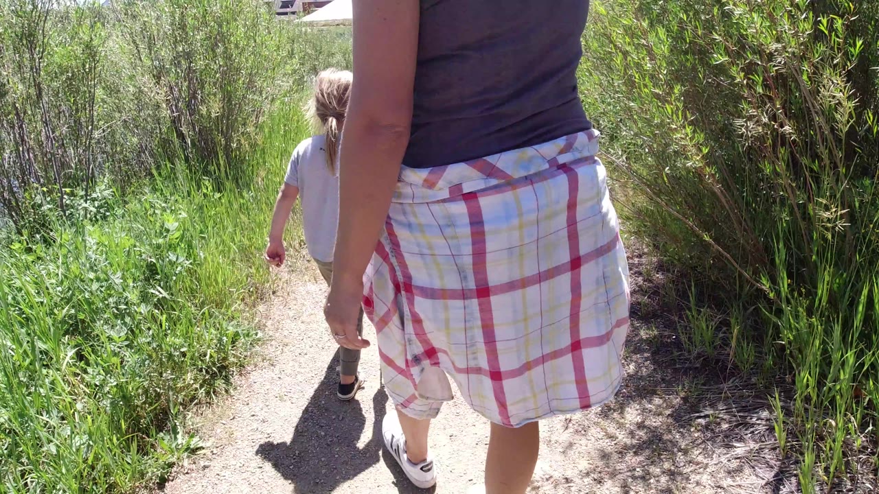 The adult remains close in the foreground while the child walks farther ahead on the dirt path through green vegetation in Winter Park, Colorado.