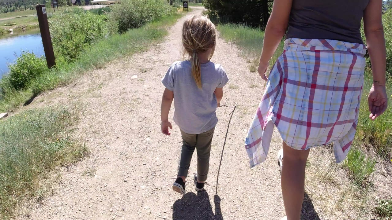 Adult and child walking away on a dirt path beside grass and water.