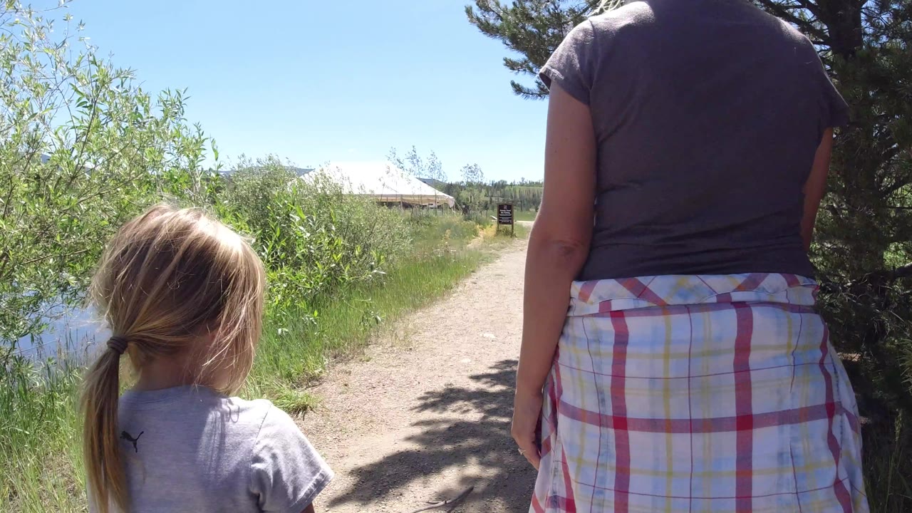 The woman stays centered while the child walks slightly left on the Winter Park path, with bright sky, roadside greenery, and a round white tent-like structure ahead.