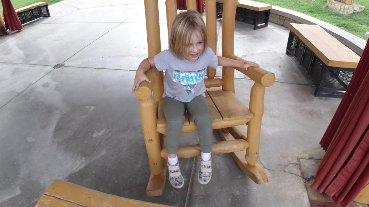 At 0:00, a young girl sits facing the camera in a large wooden rocking chair in Winter Park, Colorado, with red curtains and outdoor benches around the covered patio.
