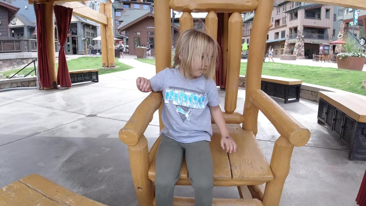 Blonde child seated on a large wooden rocking chair in a public square.