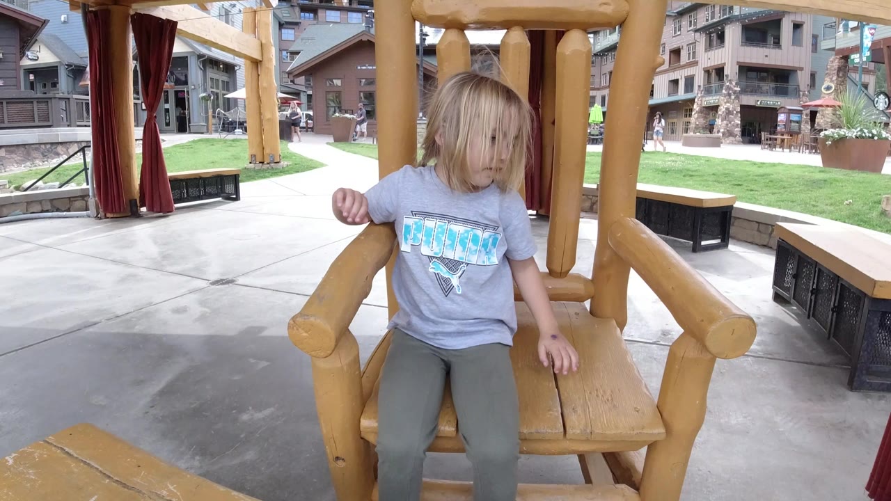 The child looks down toward the chair seat in Winter Park, Colorado, with the chair arms close to camera and the square's walkways and buildings behind.