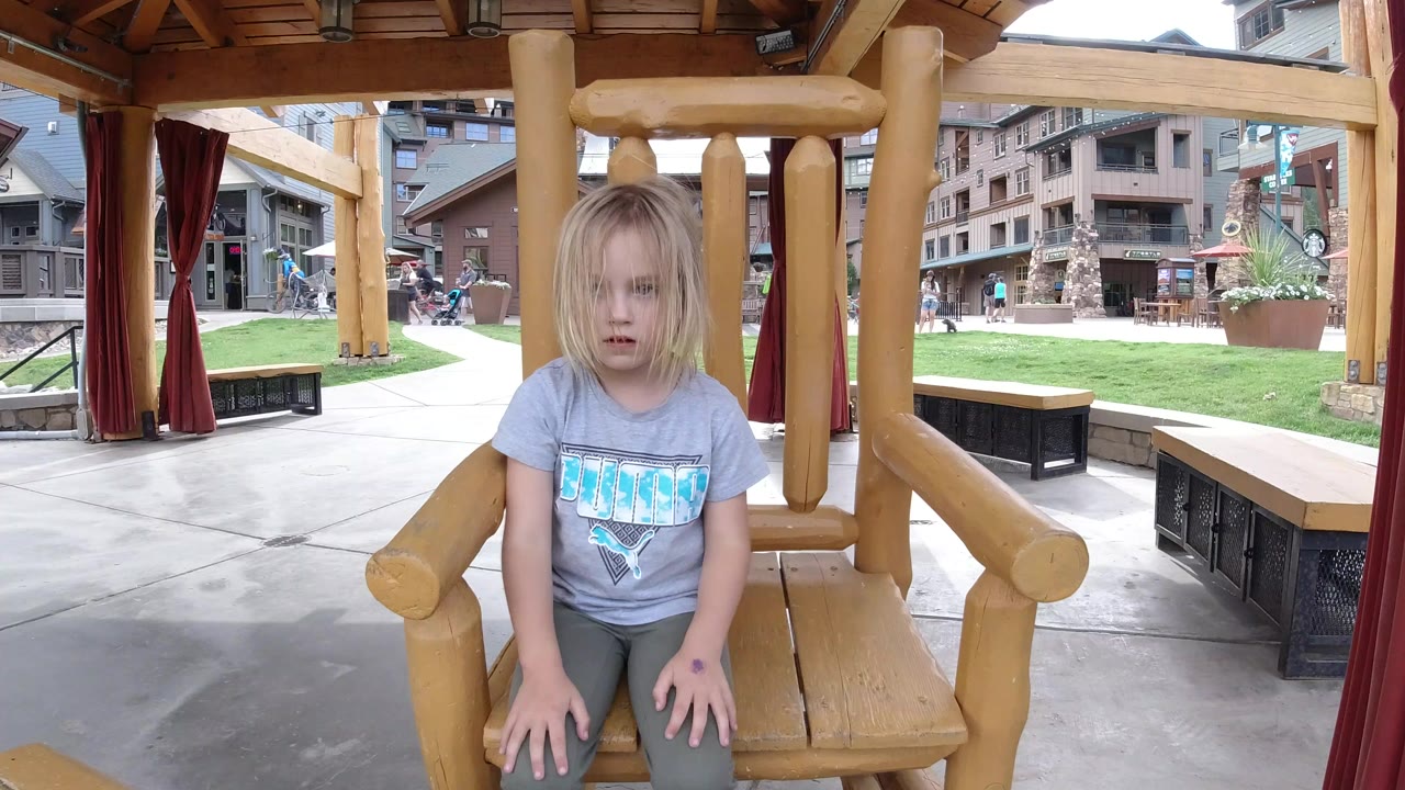 Young girl seated on a large wooden rocking chair under a shelter.