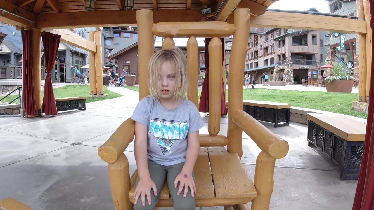 The child remains seated near the front of the large wooden chair, looking toward the camera as the covered structure and Winter Park plaza background stay in view.