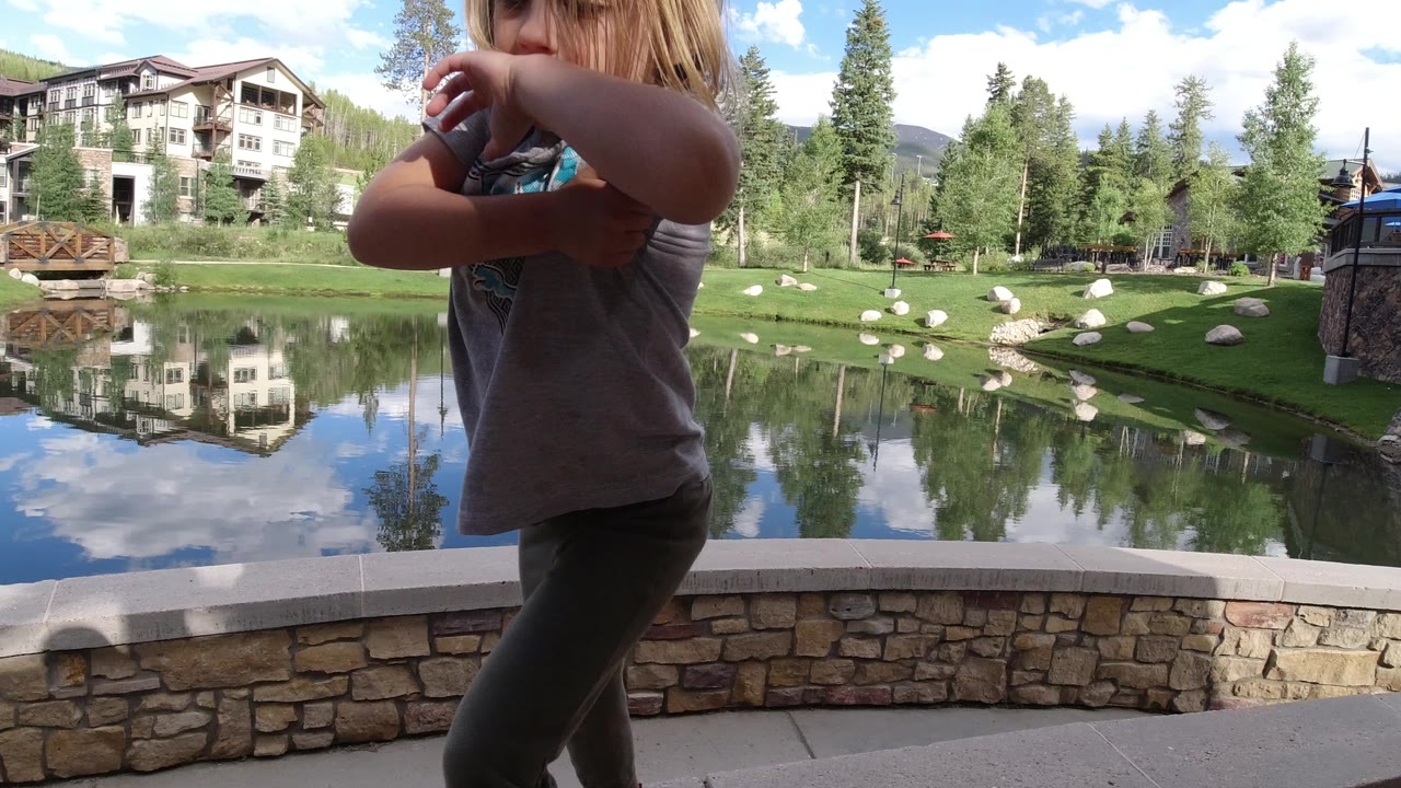 The girl moves across the near foreground beside the Winter Park lake, leaving the reflective water, green bank, conifers, birds, and lodge-style buildings visible behind her.