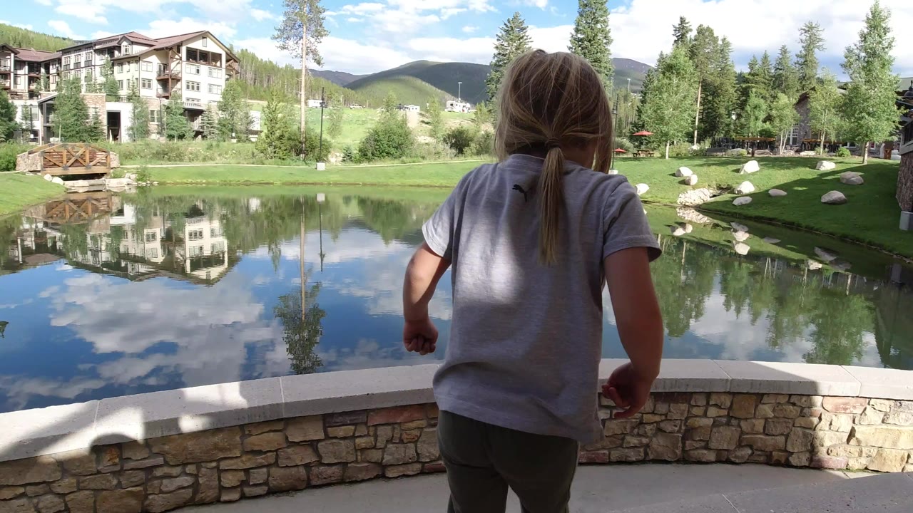 A young girl in a gray shirt is seen from behind near a low stone wall in Winter Park, Colorado, with a reflective pond, lodge buildings, trees, and hills beyond her.