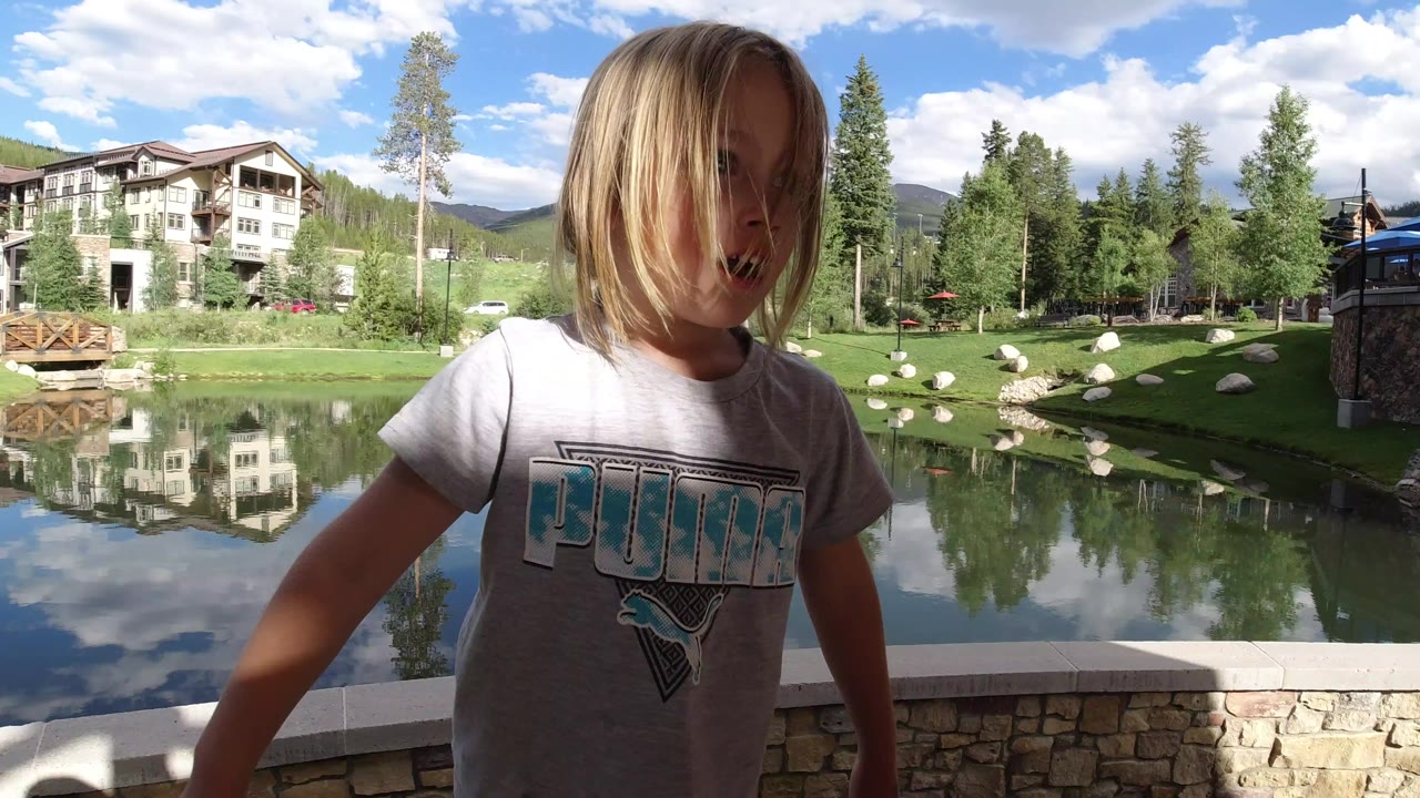 Blonde girl close to camera beside a pond and lodge buildings.
