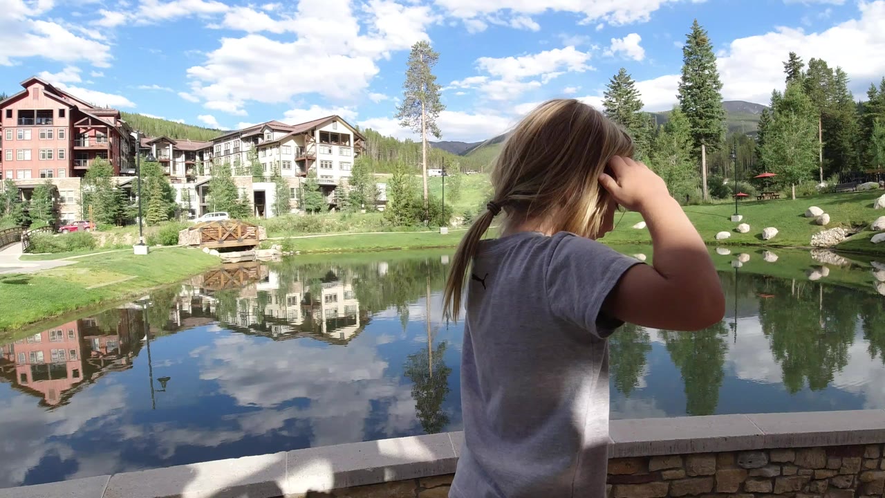 A blonde child stands with their back partly to camera beside a stone wall, looking toward a reflective lake and lodge-style buildings in Winter Park, Colorado.