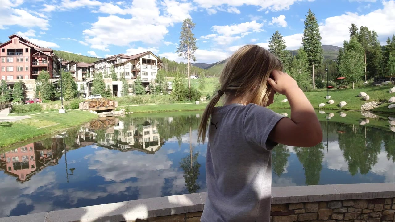 The child remains near the right edge while the lake reflects clouds, trees, and light-colored mountain buildings in Winter Park, Colorado.