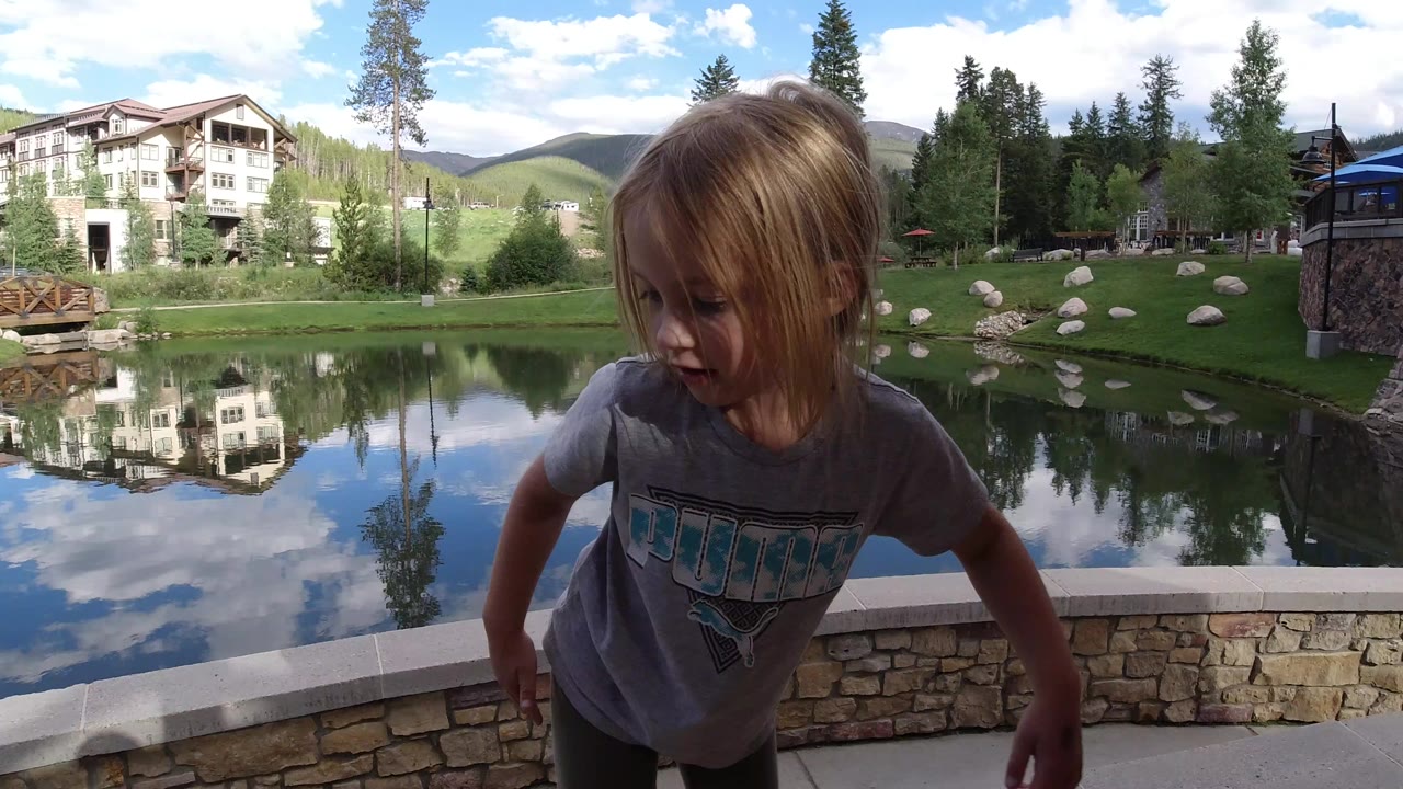 A young blond girl stands close to camera at an outdoor pond area in Winter Park, Colorado, with a stone wall, reflected building, lawn, trees, hills, and clouds behind her.