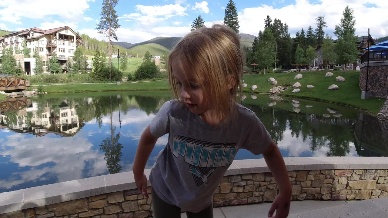 The girl remains in the foreground at Winter Park, Colorado, turned slightly downward as the pond reflects a multi-story building, trees, hills, and blue sky behind her.
