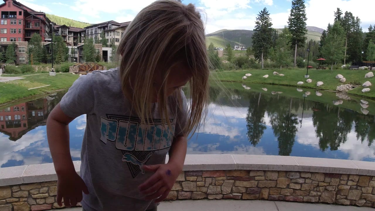 The child leans forward and looks down near the stone wall, while the lake reflects clouds, trees, and resort buildings in Winter Park, Colorado.
