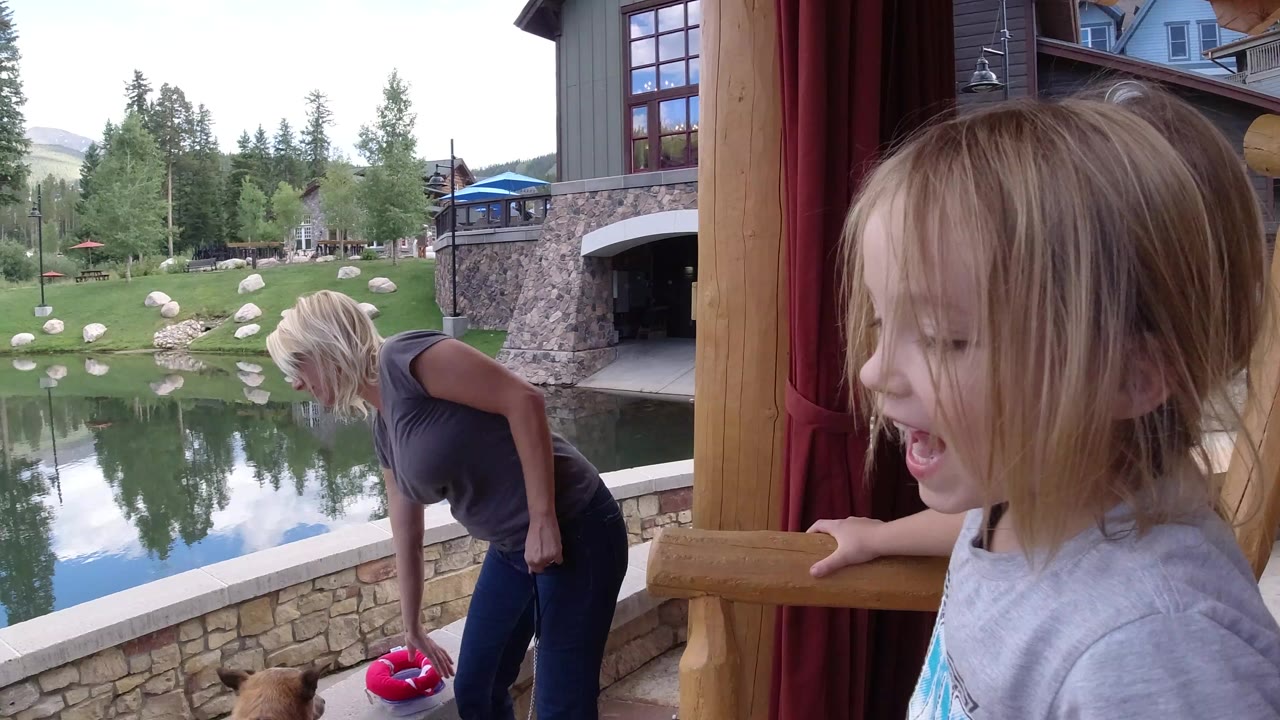 Child near an oversized wooden chair as a woman bends toward a dog by a pond.