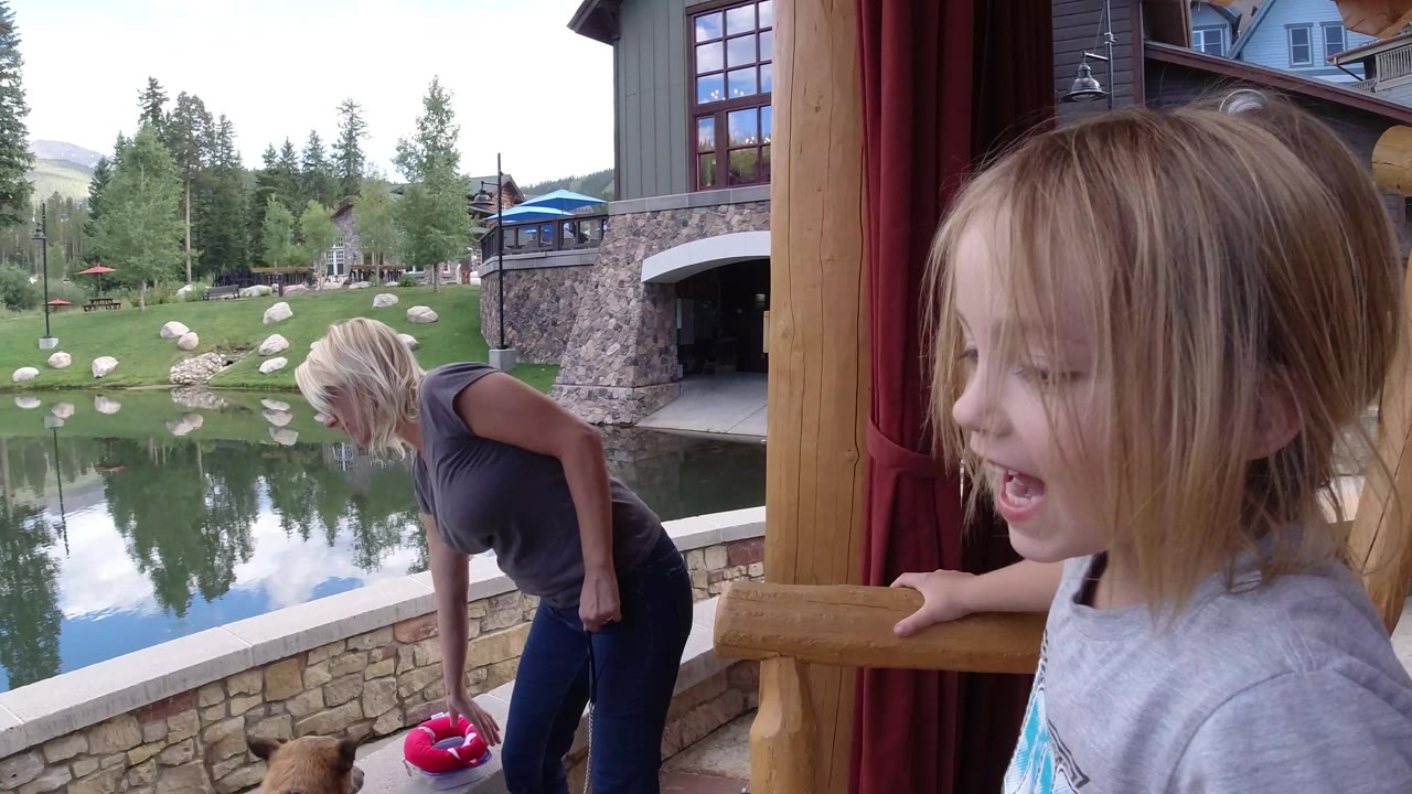 A young child remains in the foreground by the wooden chair as the woman leans over the dog, with the Winter Park pond, rocks, and trees filling the background.