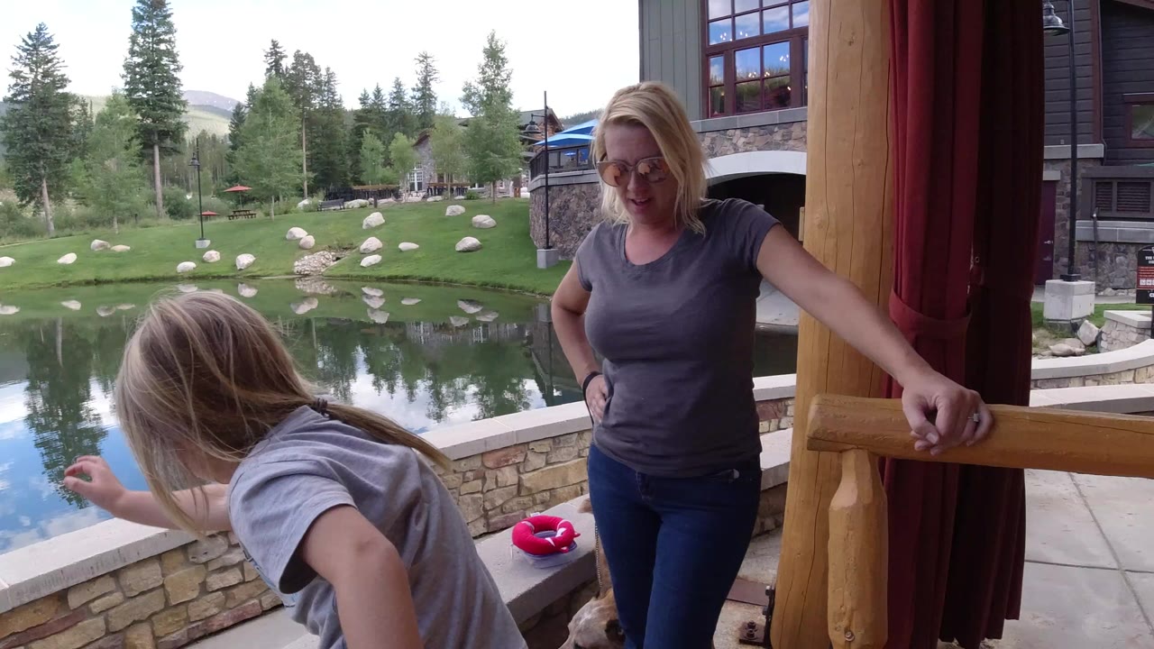 In Winter Park, Colorado, an adult woman stands by a porch post while a young girl leans forward near a stone-edged pond with trees and lodge buildings beyond.
