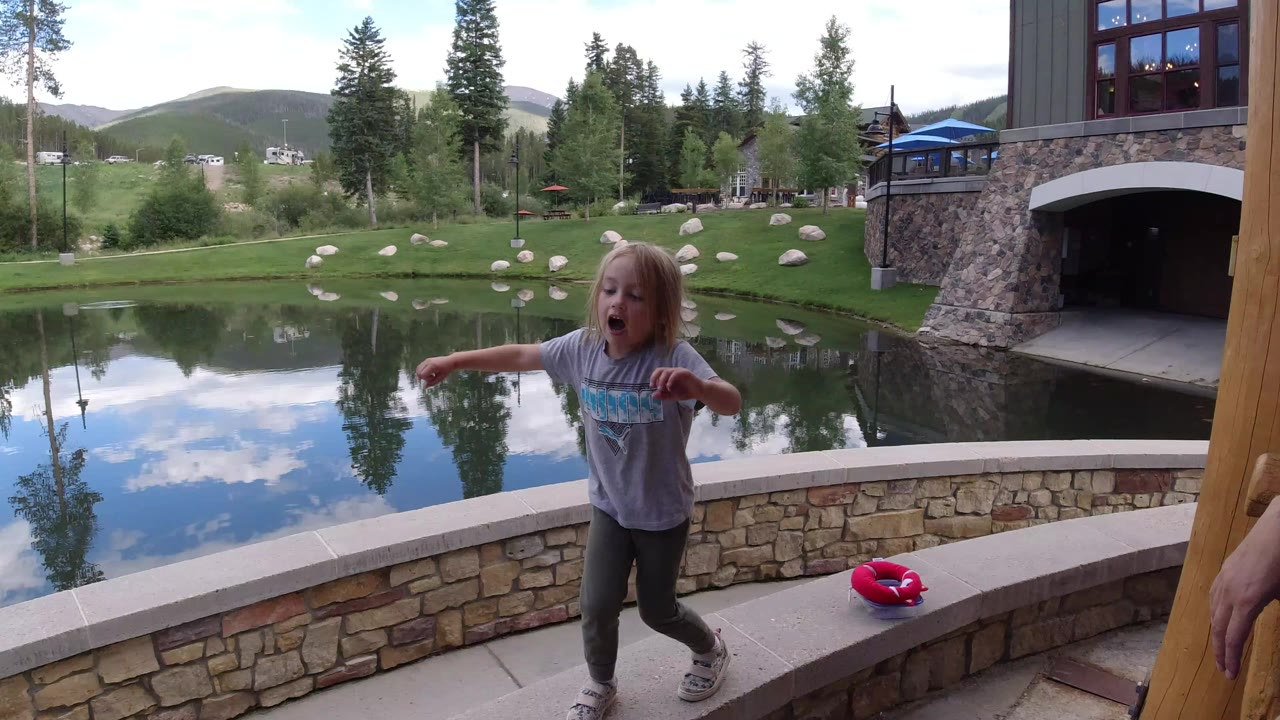 Child on a deck beside a reflective pond and mountain homes.