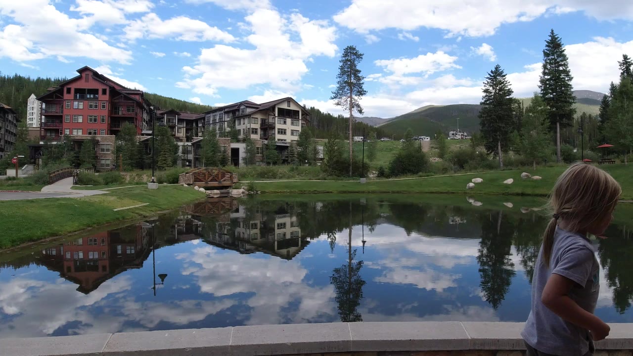 Child at a reflective mountain lake with lodge buildings and trees.