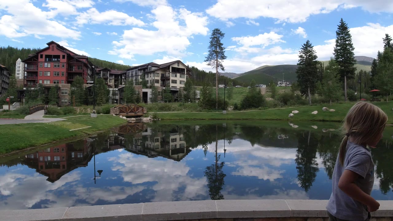 A child remains near the foreground ledge as the lake reflects clouds, blue sky, tall trees, and multi-story buildings in Winter Park, Colorado.