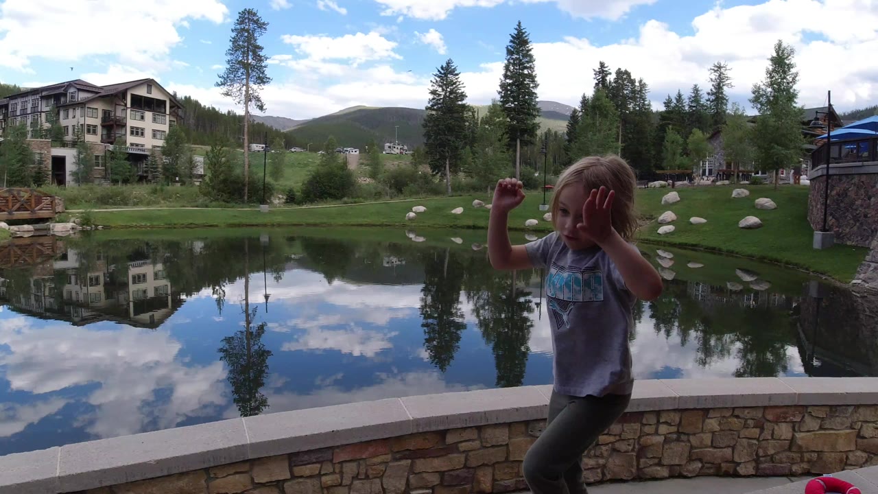 A young girl stands near the stone edge of a pond in Winter Park, Colorado, with lodge-style buildings, trees, hills, and cloud reflections behind her.