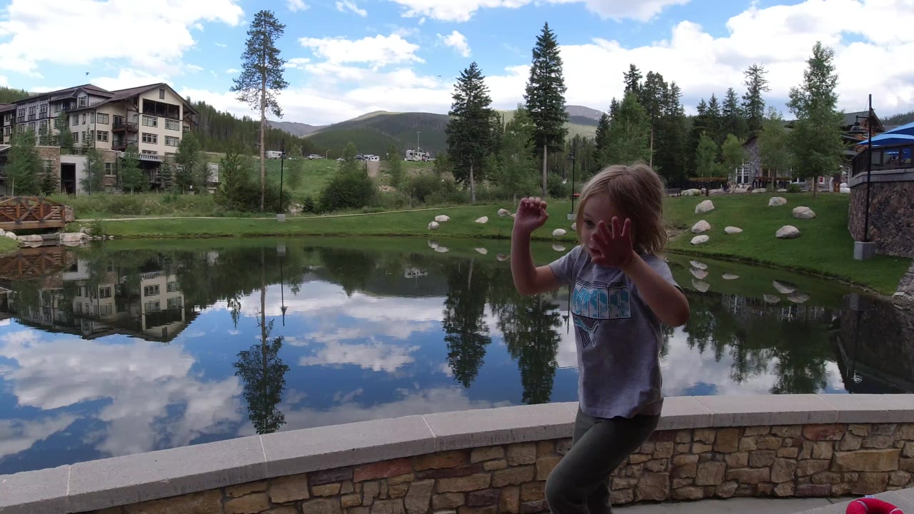 The girl raises both hands beside the pond in Winter Park, Colorado, while lodge buildings and tall trees reflect in the still water.