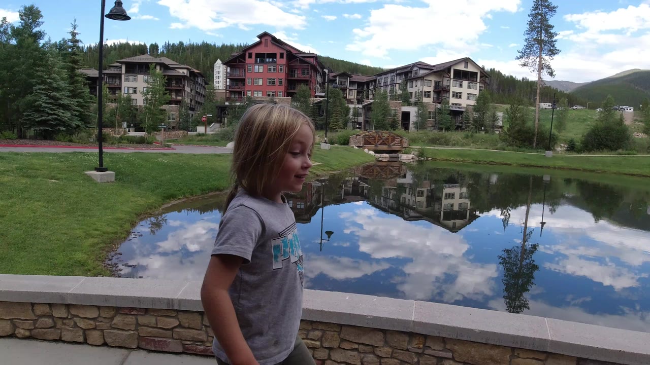 Profile view of the young girl near the stone wall in Winter Park, Colorado, with calm pond reflections, multi-story lodge buildings, lawns, and evergreen trees behind her.