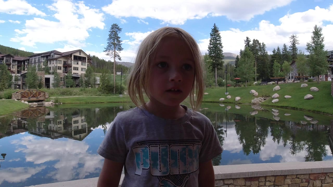 Young girl standing beside a pond with lodge buildings and trees behind her.